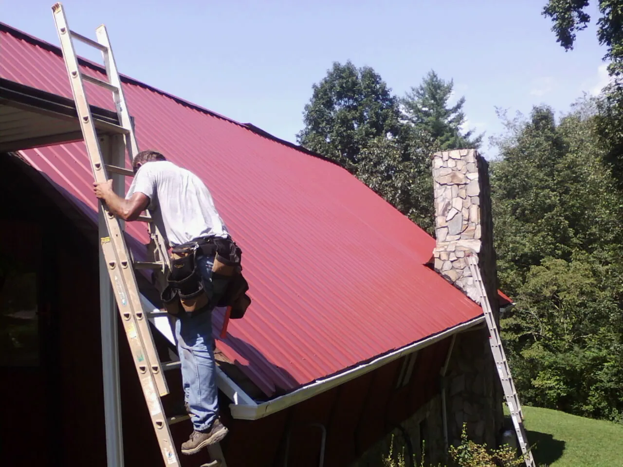 Roofer on a ladder beside a red metal roof and stone chimney — Soffit & Fascia Repair by Roofing Brothers in Lewisville